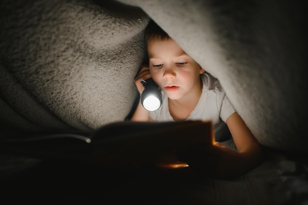 Portrait of cute little boy reading in bed avec lampe de poche dans une pièce sombre en appréciant les contes de fées