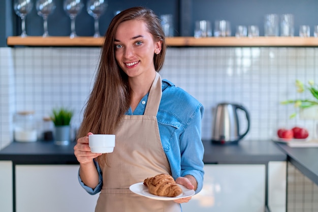 Portrait of cute young happy smiling woman baker tenant une tasse blanche et un croissant de plaque à la cuisine loft