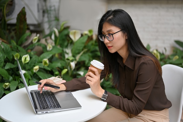 Portrait of female freelancer holding paper cup et main tapant sur ordinateur portable tout en travaillant en plein air dans le jardin