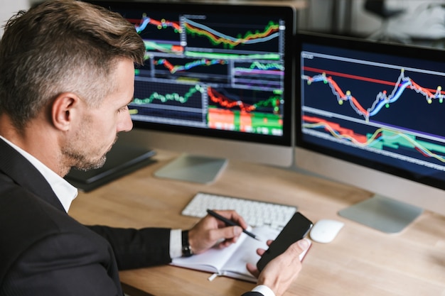 Portrait of handsome businessman 30s wearing suit using mobile phone tout en travaillant avec des graphiques numériques sur ordinateur au bureau