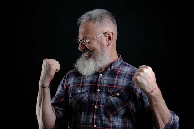 Portrait of happy guy européen mature barbu joyeux et émotif, levant les poings haut et hurlant, triomphant ou applaudissant en regardant un match de football, mur noir