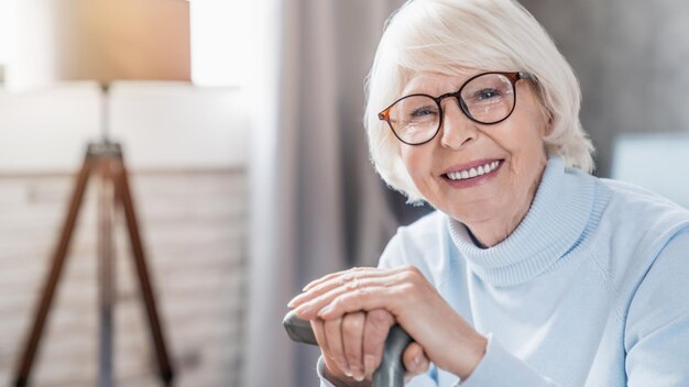 Portrait of happy mature woman in eyeglasses holding cane alors qu'il était assis sur le canapé à la maison