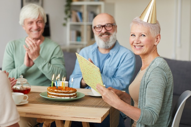 Portrait of happy senior woman reading carte de voeux et souriant à la caméra tout en célébrant l'anniversaire avec ses amis