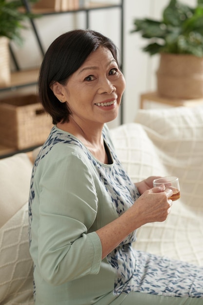 Portrait of happy senior woman sitting on sofa in living room et boire une tasse de thé