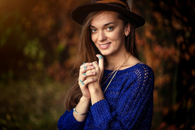 Portrait of happy smiling boho chic femme à la mode en chapeau et pull bleu tricoté avec collier portant des bagues en argent avec pierre turquoise dans la forêt d'automne. Fille de bijoux élégant avec mode boho