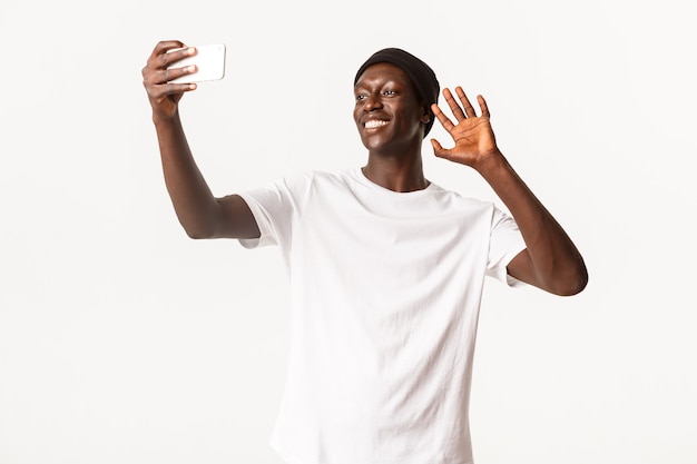 Portrait of happy smiling guy afro-américain parler avec des amis via appel vidéo, tenant le smartphone et agitant la main en geste bonjour