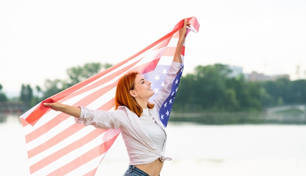 Portrait of happy smiling red haired girl holding USA drapeau national dans ses mains. Jeune femme positive célébrant la fête de l'indépendance des États-Unis.