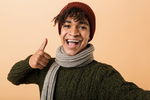 Portrait of happy teen man wearing hat et écharpe gesticulant, isolé sur mur jaune