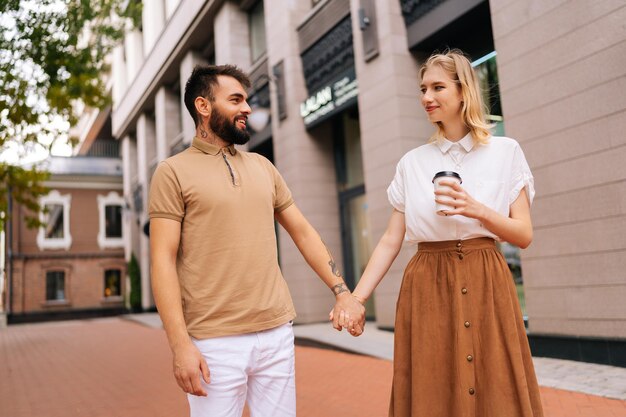 Portrait of happy young couple in love standing holding hands on city street and looking on each Bel homme barbu et jolie femme blonde marchant en buvant