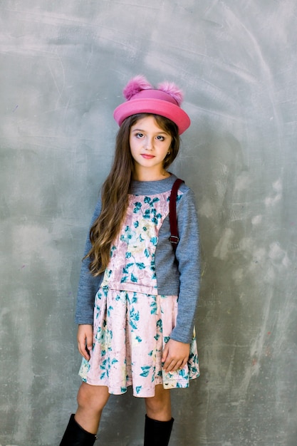 Portrait of little fashion kid girl in pink cap and dress with flowers and a brown backpack on a gray wall