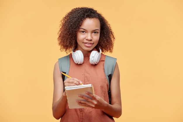 Portrait of smiling curieux étudiant afro-américain fille aux cheveux bouclés portant des écouteurs sans fil prendre des notes dans le bloc-notes sur fond jaune