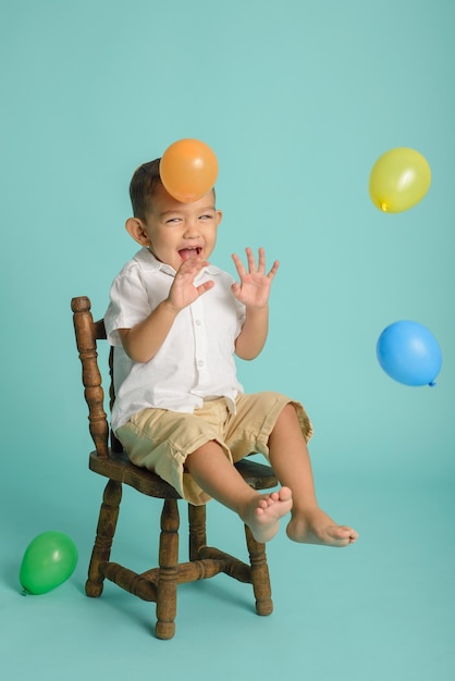 Portrait of smiling little boy habillé en blanc jouant avec des ballons assis sur une chaise en bois fond bleu