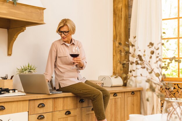 Portrait of smiling senior woman holding verre de vin tout en utilisant un ordinateur portable sur la table de la cuisine. Concept de travail indépendant à domicile.