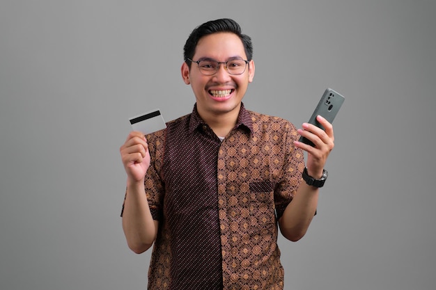 Portrait of smiling young Asian man wearing batik shirt holding carte de crédit et smartphone isolé sur fond gris