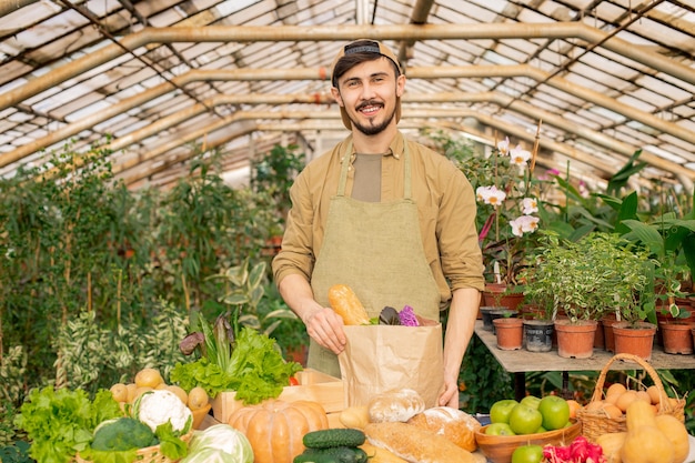 Portrait of smiling young farmer barbu en cap et tablier debout au comptoir avec divers produits et emballage de légumes frais dans un sac en papier