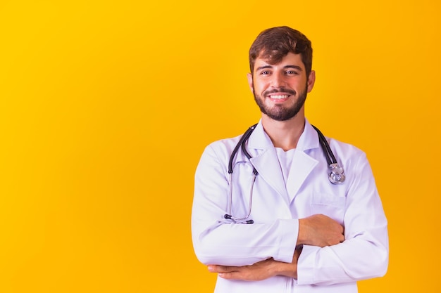 Portrait of smiling young male doctor avec stéthoscope autour du cou debout avec les bras croisés en blouse blanche isolé sur fond jaune