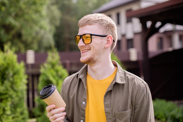 Portrait of smiling young mec barbu dans des verres jaunes habillés avec désinvolture tenant une tasse de papier jetable à l'extérieur par beau jour d'été