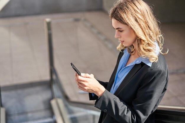 Portrait of young businesswoman on escalator using smartphone and smiling tap on mobile phone wearin