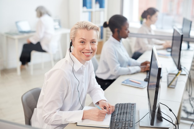 Portrait d'opératrice portant un casque et souriant alors qu'il était assis en ligne dans le centre d'appels ou l'intérieur du bureau