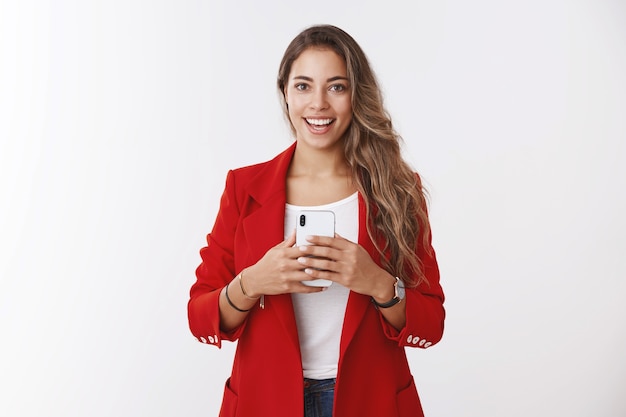 Portrait optimiste charismatique belle femme aux cheveux bouclés tenant un smartphone souriant prenant selfie miroir, employé smm indépendant capturant une photo en ligne sur les médias sociaux, mur blanc