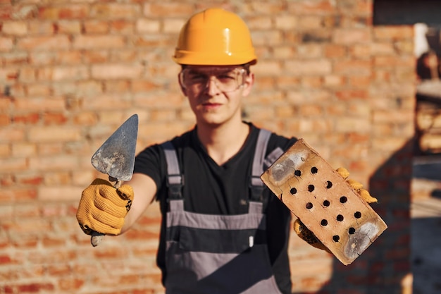 Portrait d'ouvrier du bâtiment en uniforme et équipement de sécurité qui se tient debout sur la construction et la tenue de briques et d'outils