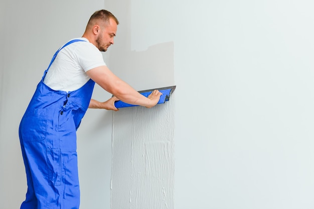 Portrait d'un ouvrier en salopette et tenant un couteau à mastic dans ses mains sur fond de mur plâtré. Concept de travaux de réparation et de construction