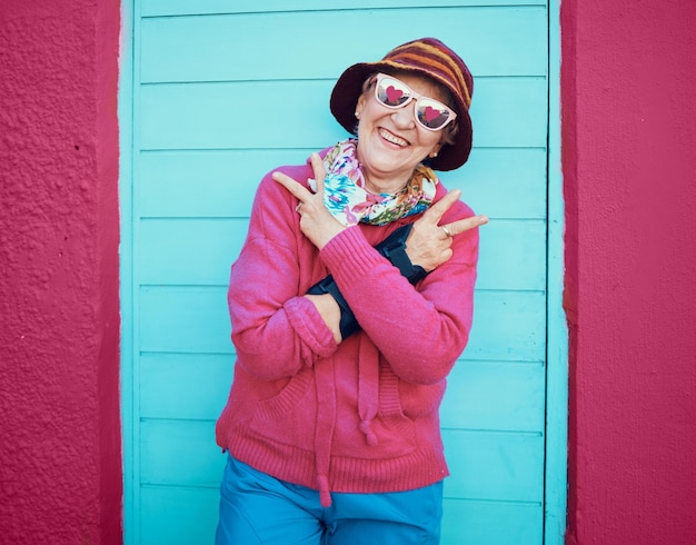 Photo portrait paix et funky avec une femme senior en plein air debout contre une porte bleue et un fond de mur rouge lunettes mains et hip hop avec une femme mature heureuse faisant un signe de la main ou un geste à l'extérieur