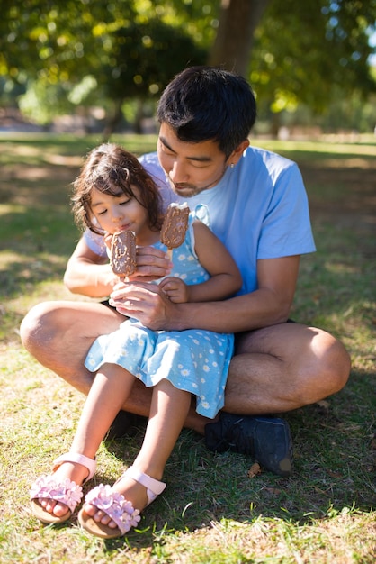 Portrait de papa asiatique nourrissant la crème glacée de sa fille dans le parc. Père prudent assis sur l'herbe verte avec un petit enfant sur ses genoux lui donnant de la crème glacée. Moments heureux ensemble, loisirs, concept de parentalité