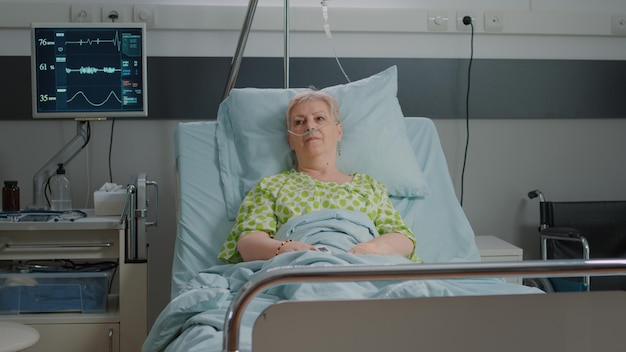 Portrait d'un patient âgé allongé dans un lit d'hôpital avec tube d'oxygène nasal et sac d'égouttement IV pour se remettre d'une maladie. Femme connectée à un moniteur de fréquence cardiaque, en attente d'une assistance médicale.