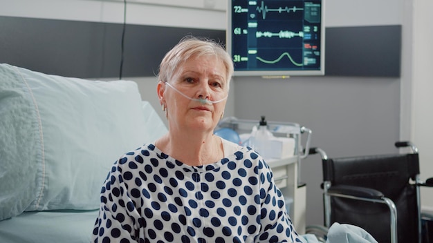 Portrait d'un patient malade assis dans un lit d'hôpital avec un tube d'oxygène nasal et un sac d'égouttement IV pour la récupération médicale. Senior woman with disease et oxymètre connecté au moniteur de fréquence cardiaque