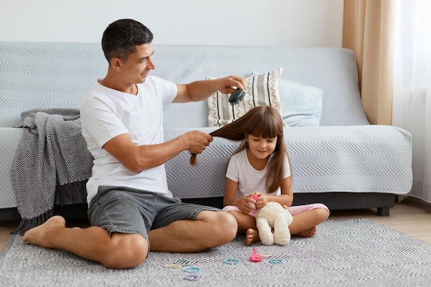 Portrait d'un père attentionné peignant les cheveux pour son enfant mignon homme portant un t-shirt blanc de style décontracté et un jean court assis sur le sol avec son enfant passant du temps ensemble