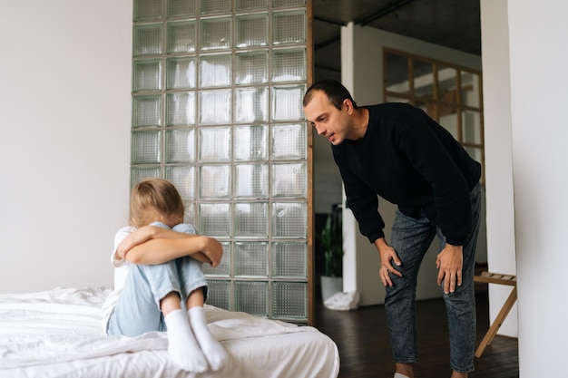 Portrait d'un père en colère grondant élevant la voix en criant à la triste petite fille difficile têtue assise sur le lit serrant le genou pleurant couvrant le visage