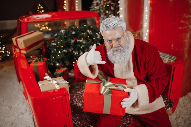 Portrait d'un père Noël élégant avec un cadeau dans ses mains, beaucoup de cadeaux à l'arrière d'une voiture. Montre le signe de l'appel, téléphone. Préparer des cadeaux pour la célébration de Noël et du nouvel an