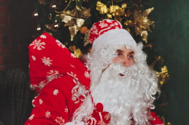 Portrait d'un père Noël émotionnel avec sac de cadeaux par cheminée de Noël dans un salon décoré avec arbre de Noël. Le Père Noël vient et apporte des cadeaux la nuit. Espace de copie pour le site