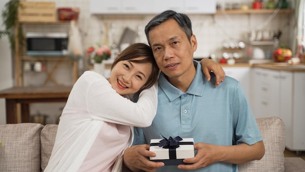 portrait d'un père retraité asiatique et d'une fille adulte regardant la caméra avec le cadeau de la fête des pères dans le salon à la maison. la femme souriante embrasse l'homme et s'appuie sur son épaule