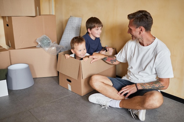 Portrait d'un père tatoué souriant jouant avec deux fils dans des boîtes en carton pendant que la famille déménage dans de nouveaux