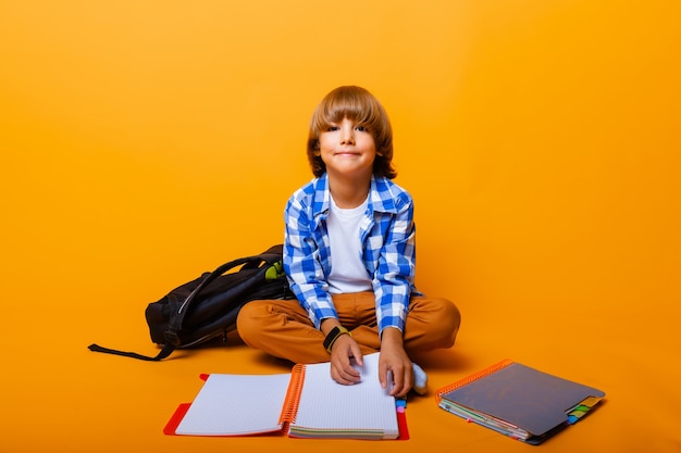 Portrait d'un petit écolier avec un livre sur fond jaune, studio. écolier assis par terre dans le studio
