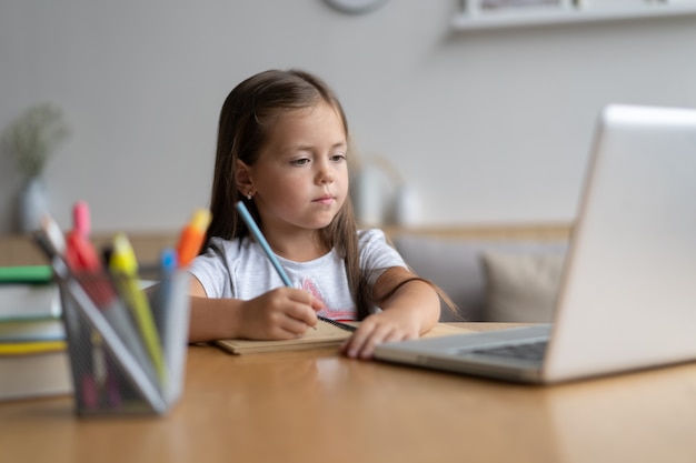 Portrait d'un petit élève heureux qui apprend à la maison. Petite fille souriante appréciant de faire des cours dans le salon. Écolière intelligente regardant la caméra, étudiant à distance en ligne.