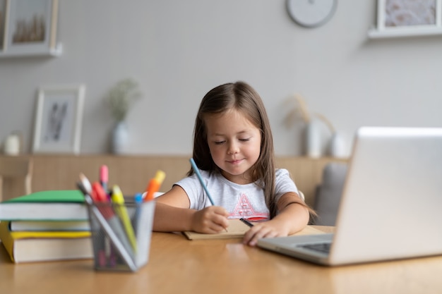 Portrait d'un petit élève heureux qui apprend à la maison. Petite fille souriante appréciant de faire des cours dans le salon. Écolière intelligente regardant la caméra, étudiant à distance en ligne.