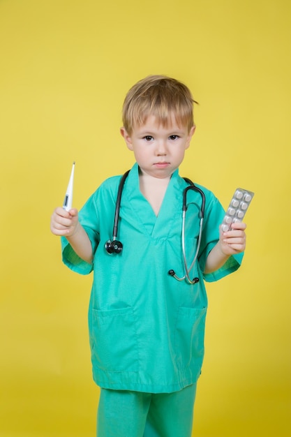 Portrait d'un petit enfant caucasien vêtu d'un manteau vert de médecins contenant des cloques avec des pilules à la main sur fond jaune