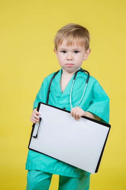 Portrait d'un petit enfant caucasien vêtu d'un manteau vert de médecins tenant une tablette à la main isolée sur jaune