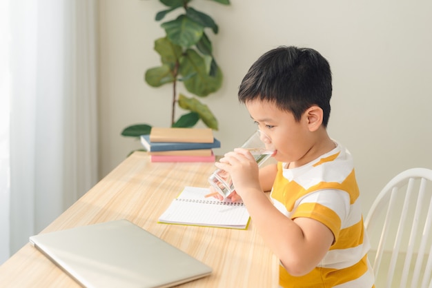 Portrait d'un petit garçon asiatique buvant de l'eau tout en utilisant un ordinateur portable, assis au bureau et faisant ses devoirs. Concept d'apprentissage et d'éducation en ligne.