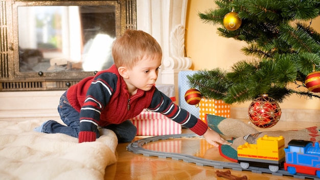 Portrait de petit garçon assis sur le sol au salon et regardant petit train sur les chemins de fer sous l'arbre de Noël