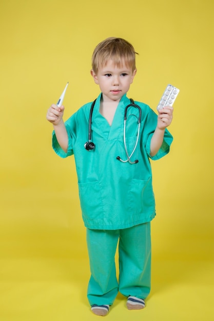 Photo portrait d'un petit garçon caucasien vêtu d'un manteau vert de médecins contenant des cloques avec des pilules à la main sur fond jaune