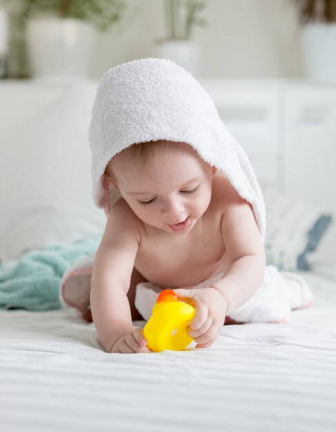 Portrait d'un petit garçon heureux dans une serviette à capuchon jouant avec un canard en caoutchouc jaune sur le lit après le bain