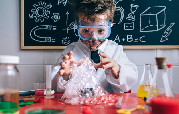 Portrait d'un petit garçon scientifique heureux avec des lunettes et un visage sale ouvrant les bras derrière du verre avec de la mousse de savon