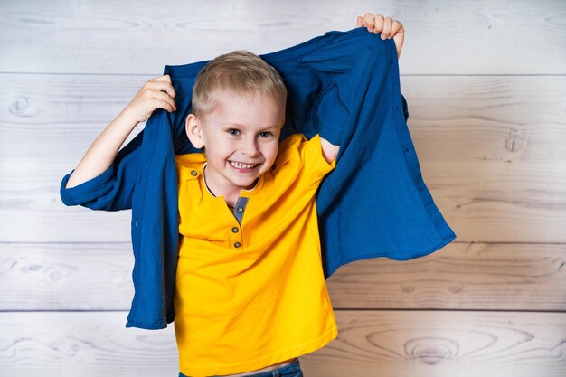 Portrait d'un petit garçon souriant en t-shirt jaune prenant sa chemise bleue sur le fond en bois