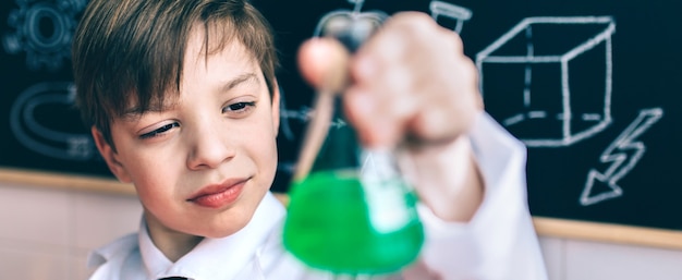 Portrait d'un petit scientifique concentré à la fiole avec un liquide vert chimique contre un tableau noir dessiné