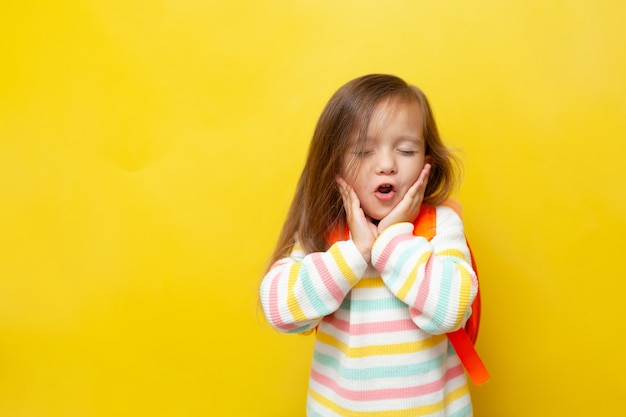 Portrait d'une petite écolière drôle avec sa bouche ouverte avec un sac à dos sur fond jaune