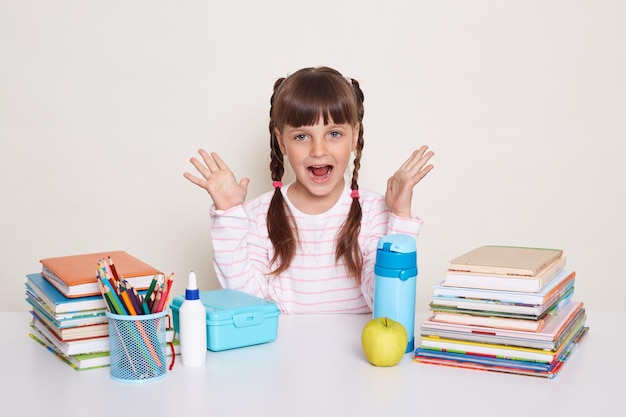 Portrait d'une petite écolière positive excitée avec des nattes portant une chemise rayée assise au bureau entourée de livres et d'autres fournitures scolaires bras levés et criant joyeusement